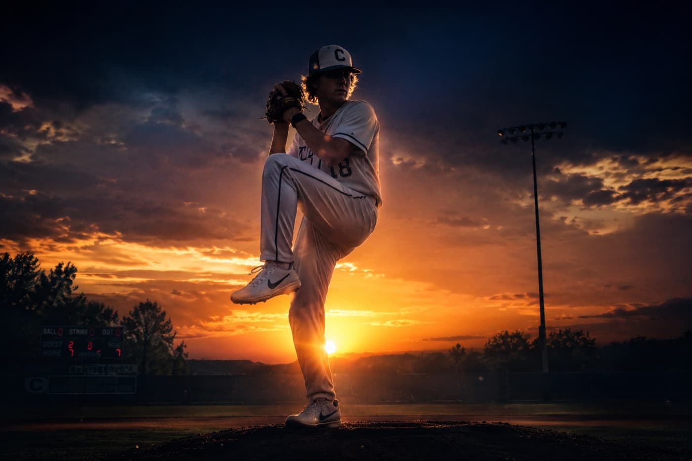 A youth pitcher mid-windup, backlit by a stadium sunset